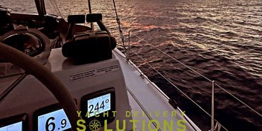 The image captures a breathtaking sunset view from the deck of a sailboat. The sky is painted with hues of orange, pink, and purple, reflecting on the calm waters of the sea. The boat's deck is visible in the foreground, with a steering wheel, a compass, and a digital display showing the heading (244°) and speed (6.9 knots). The boat's name, "YACHT DELIVERY SOLUTIONS," is engraved on the deck, suggesting that this is a professional service for delivering yachts. The overall scene evokes a sense of tranquility and adventure on the open sea.